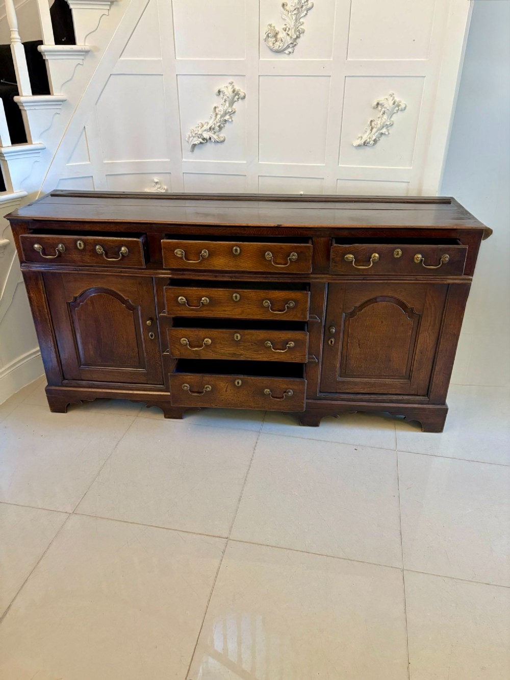 A George III dark oak dresser base featuring multiple drawers, arched cupboard doors, and brass swan-neck handles. View VII