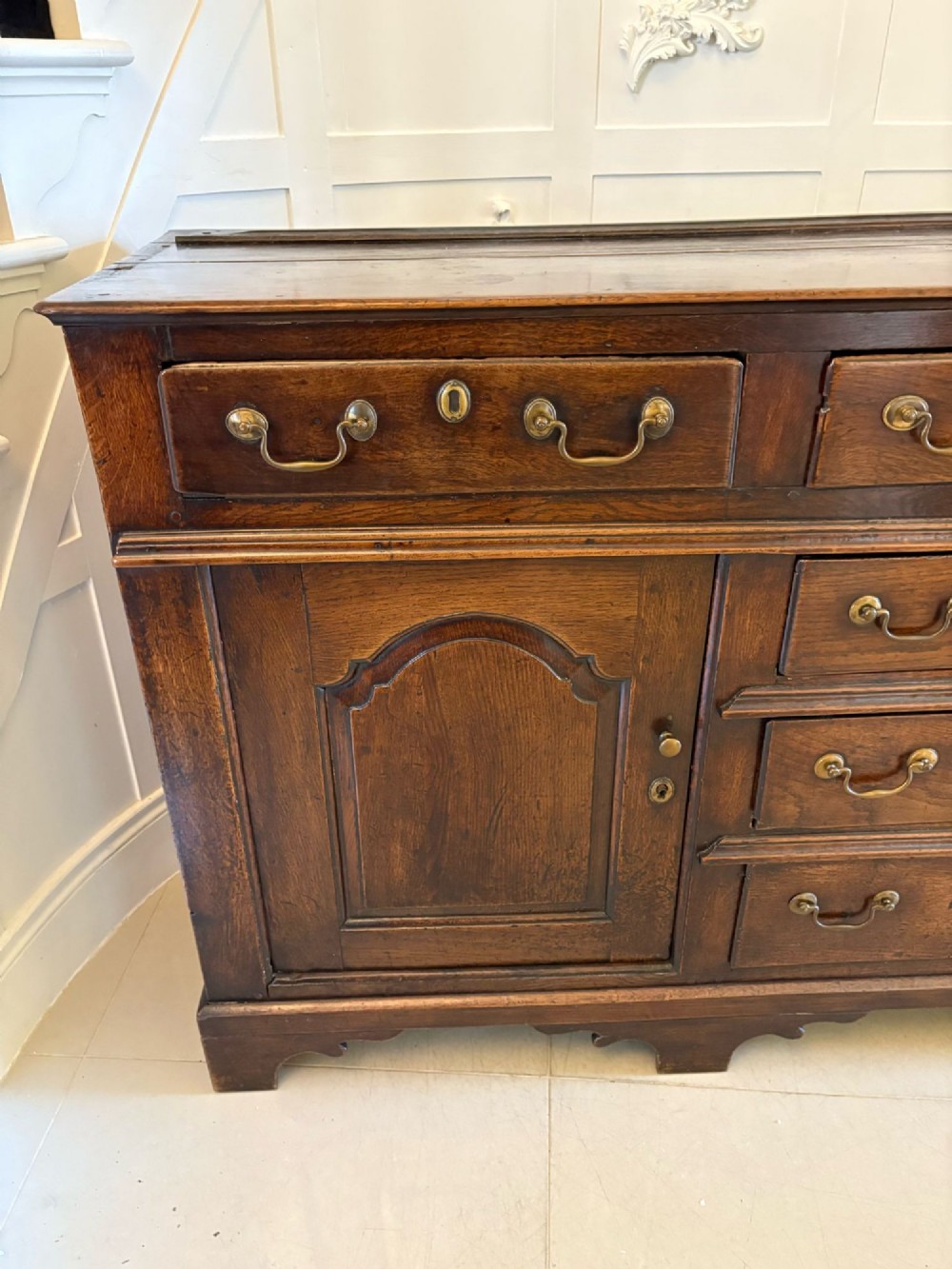 A George III dark oak dresser base featuring multiple drawers, arched cupboard doors, and brass swan-neck handles. View VIII