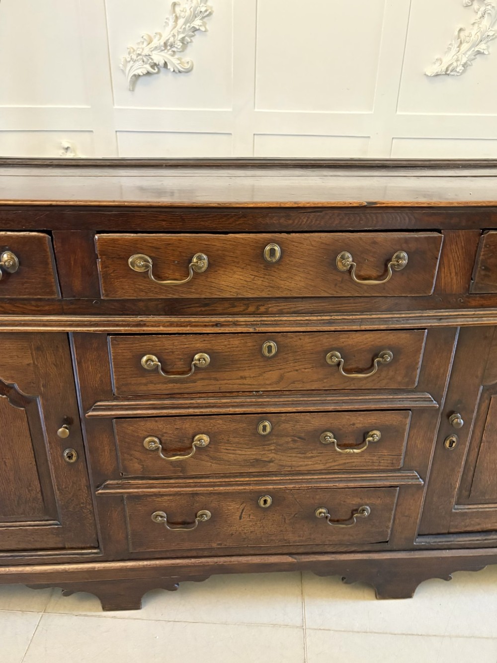 A George III dark oak dresser base featuring multiple drawers, arched cupboard doors, and brass swan-neck handles. View IX