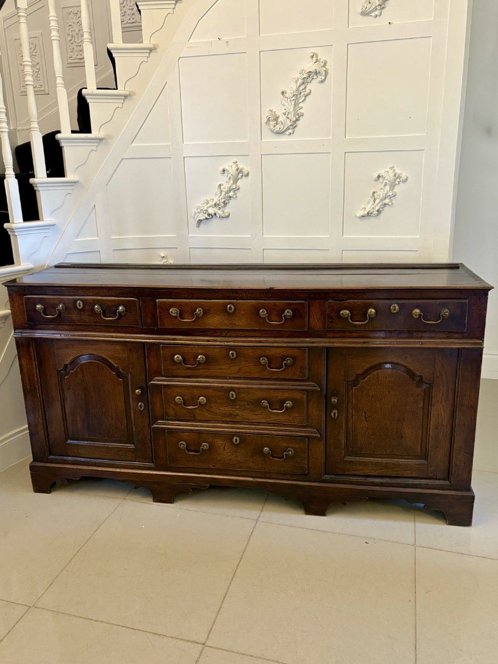 A George III dark oak dresser base featuring multiple drawers, arched cupboard doors, and brass swan-neck handles.