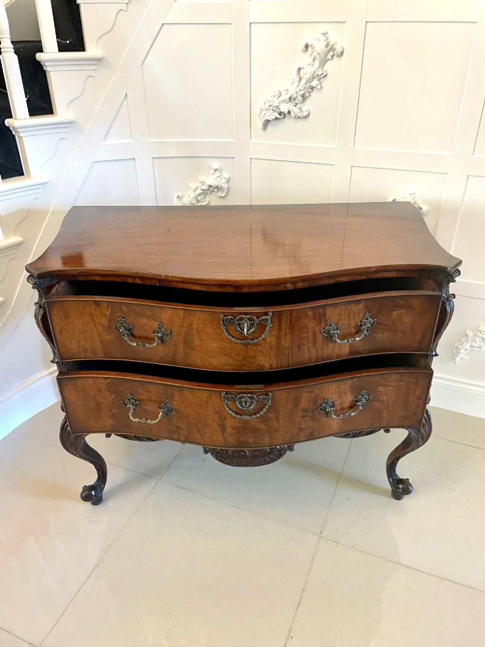 A Victorian figured mahogany serpentine chest of drawers featuring two drawers with cast brass handles, a deeply carved apron, and cabriole legs. View VII