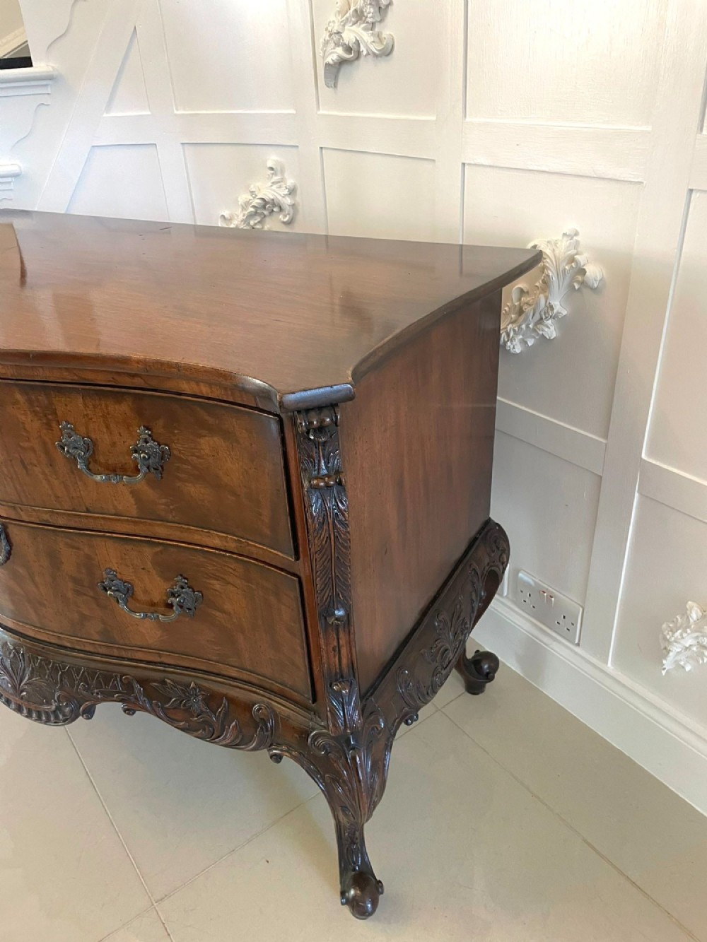 A Victorian figured mahogany serpentine chest of drawers featuring two drawers with cast brass handles, a deeply carved apron, and cabriole legs. View IX