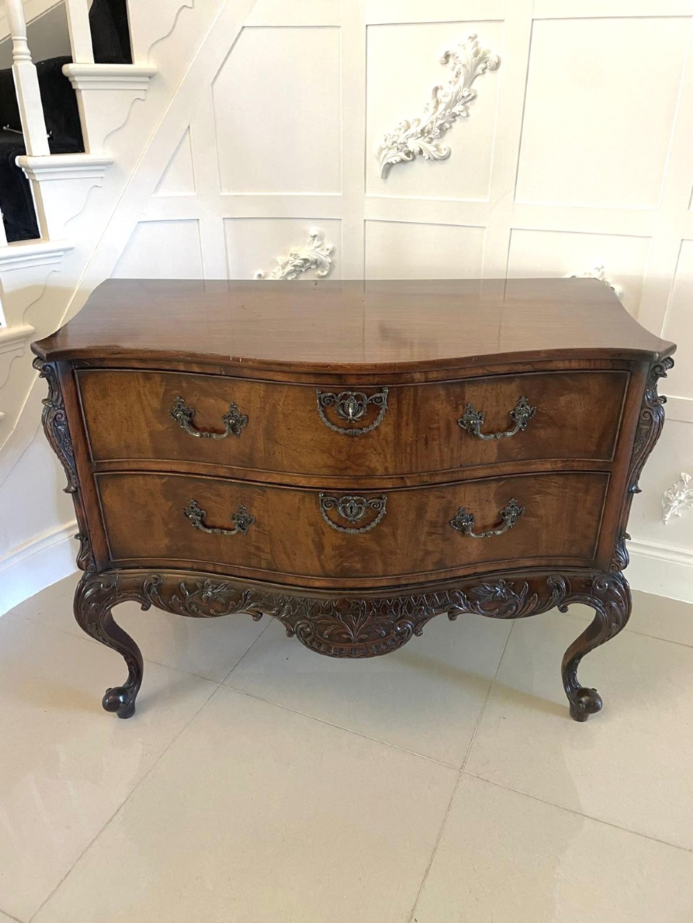 A Victorian figured mahogany serpentine chest of drawers featuring two drawers with cast brass handles, a deeply carved apron, and cabriole legs.