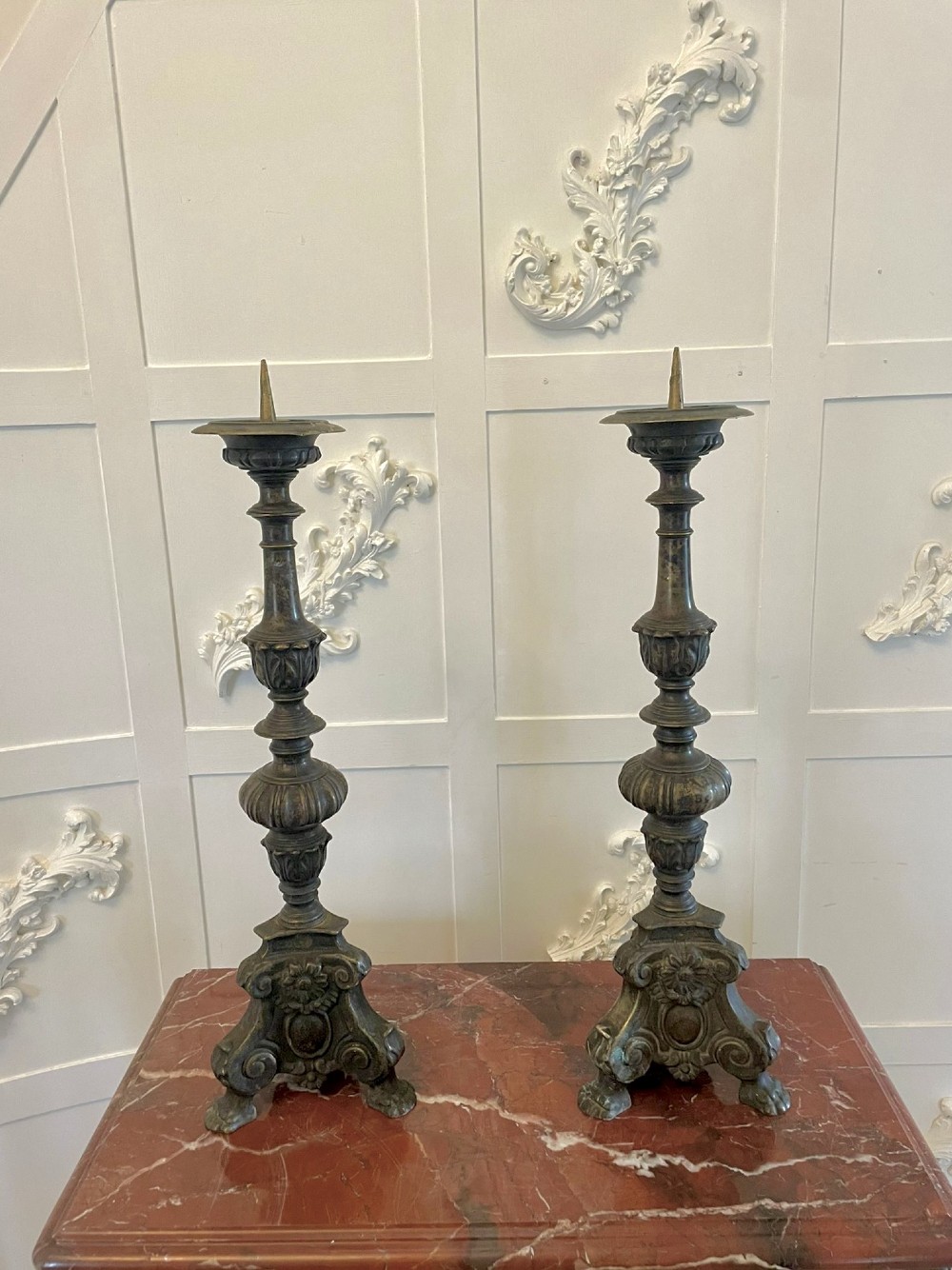 A pair of tall antique Victorian brass pricket candlesticks featuring ornate baluster shafts, foliate details, and spiked drip pans resting on decorative tripod bases.