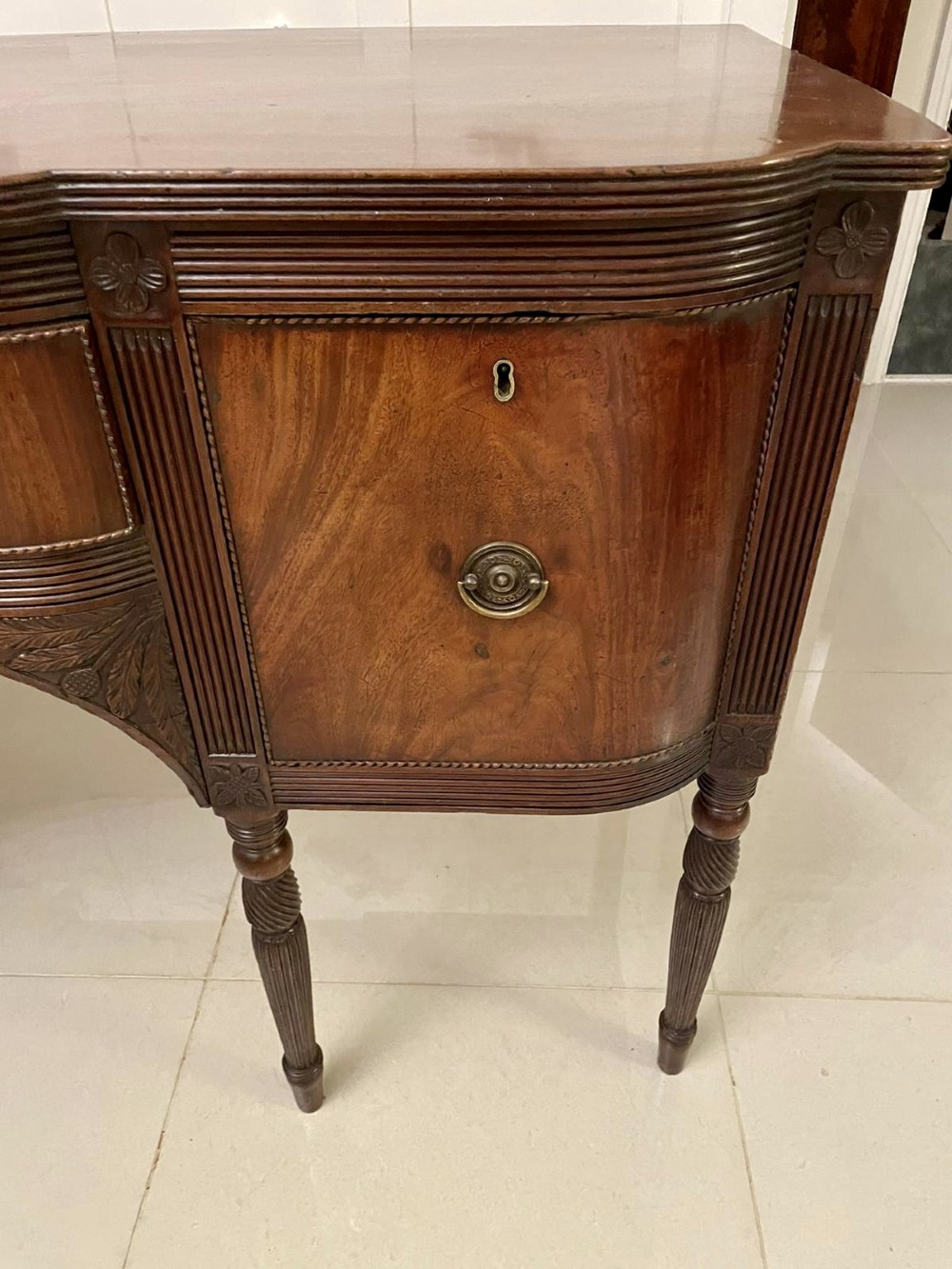 An antique George III mahogany breakfront sideboard featuring flame mahogany drawer fronts, reeded legs, and central drawer flanked by cupboards. View II