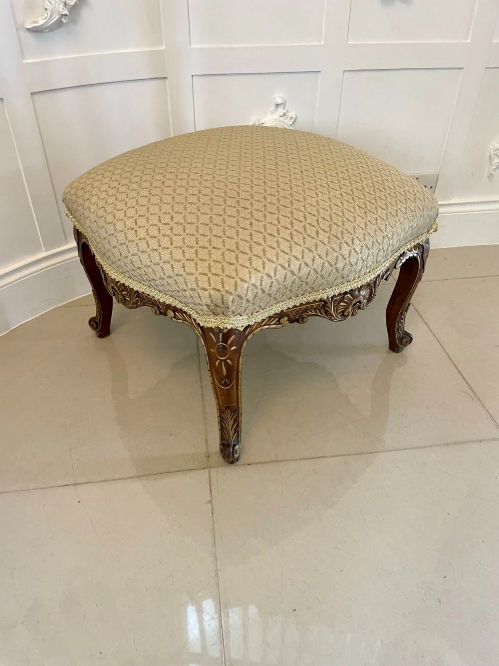 An antique Victorian low stool featuring carved walnut legs with gilt highlights and a beige geometric patterned upholstered top.
