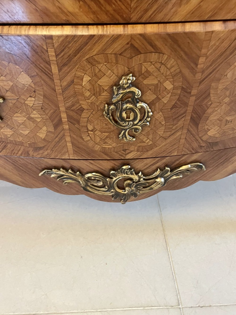 A French Louis XV style serpentine bombe commode featuring a variegated beige marble top, geometric parquetry timber veneer, three drawers, and decorative cast brass mounts. View II