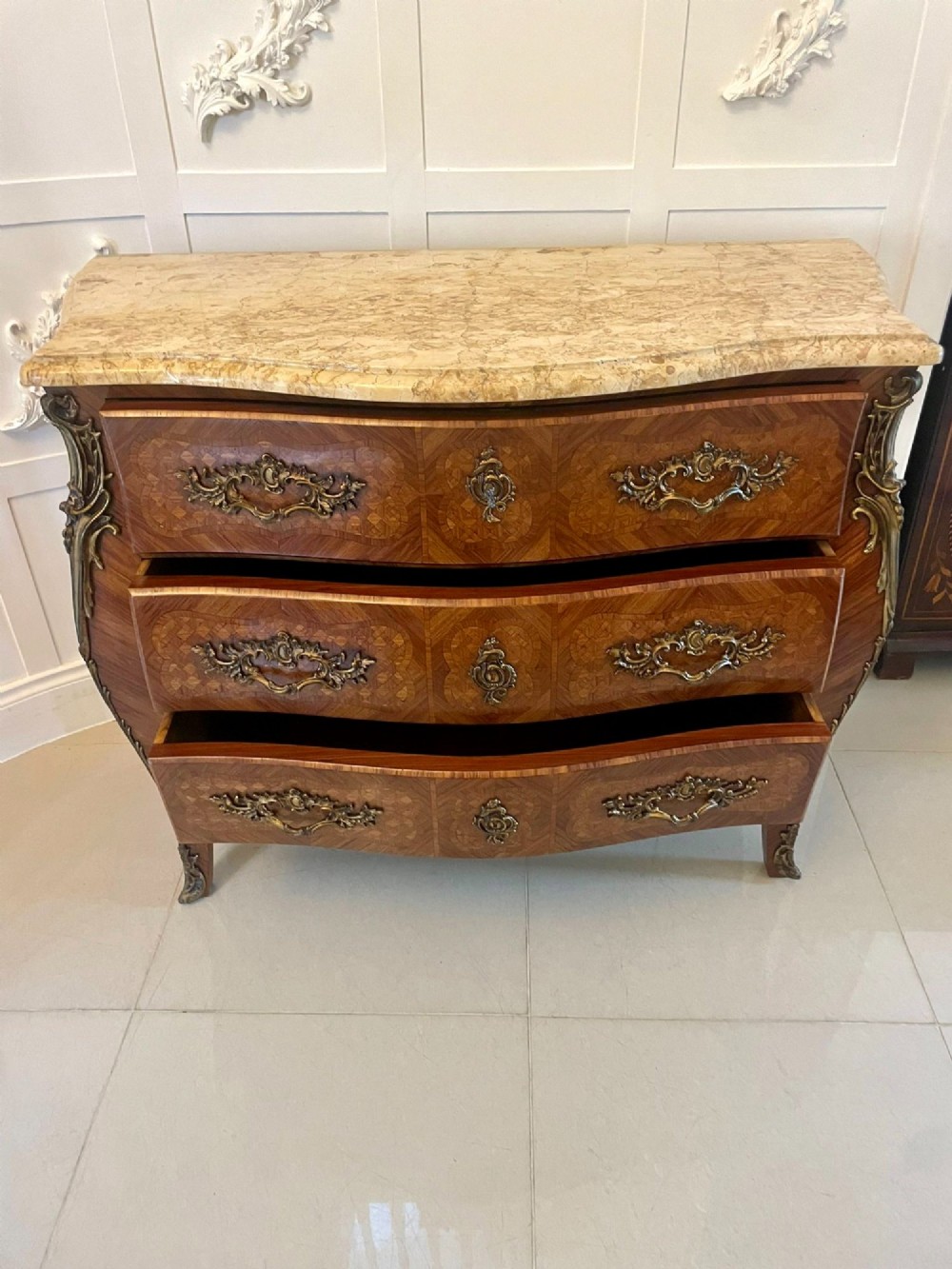 A French Louis XV style serpentine bombe commode featuring a variegated beige marble top, geometric parquetry timber veneer, three drawers, and decorative cast brass mounts. View III
