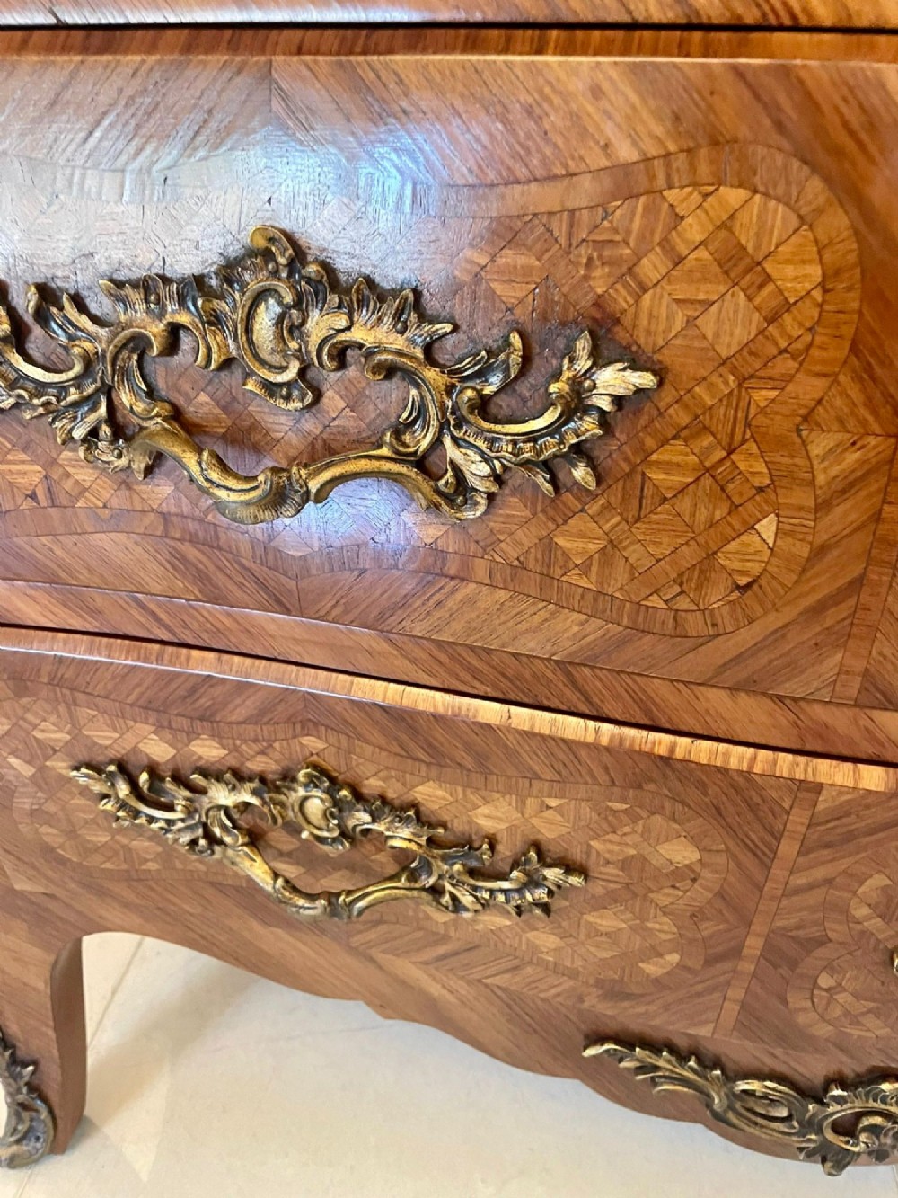 A French Louis XV style serpentine bombe commode featuring a variegated beige marble top, geometric parquetry timber veneer, three drawers, and decorative cast brass mounts. View VI