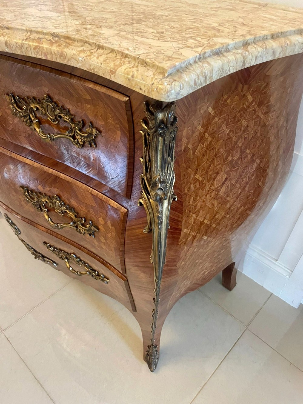 A French Louis XV style serpentine bombe commode featuring a variegated beige marble top, geometric parquetry timber veneer, three drawers, and decorative cast brass mounts. View VII