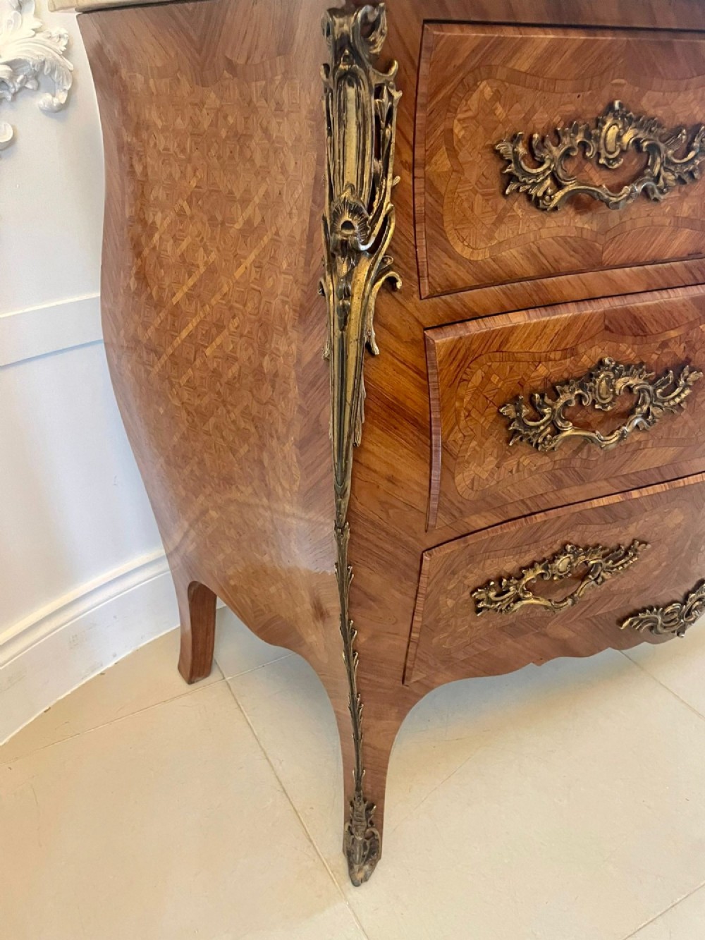 A French Louis XV style serpentine bombe commode featuring a variegated beige marble top, geometric parquetry timber veneer, three drawers, and decorative cast brass mounts. View IX