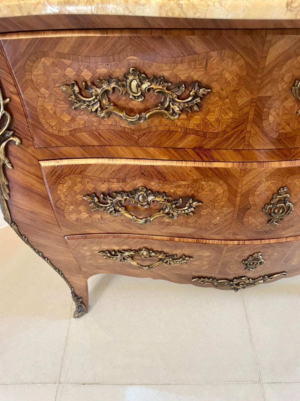 A French Louis XV style serpentine bombe commode featuring a variegated beige marble top, geometric parquetry timber veneer, three drawers, and decorative cast brass mounts. View X