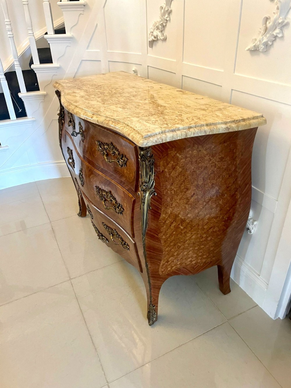 A French Louis XV style serpentine bombe commode featuring a variegated beige marble top, geometric parquetry timber veneer, three drawers, and decorative cast brass mounts. View XI