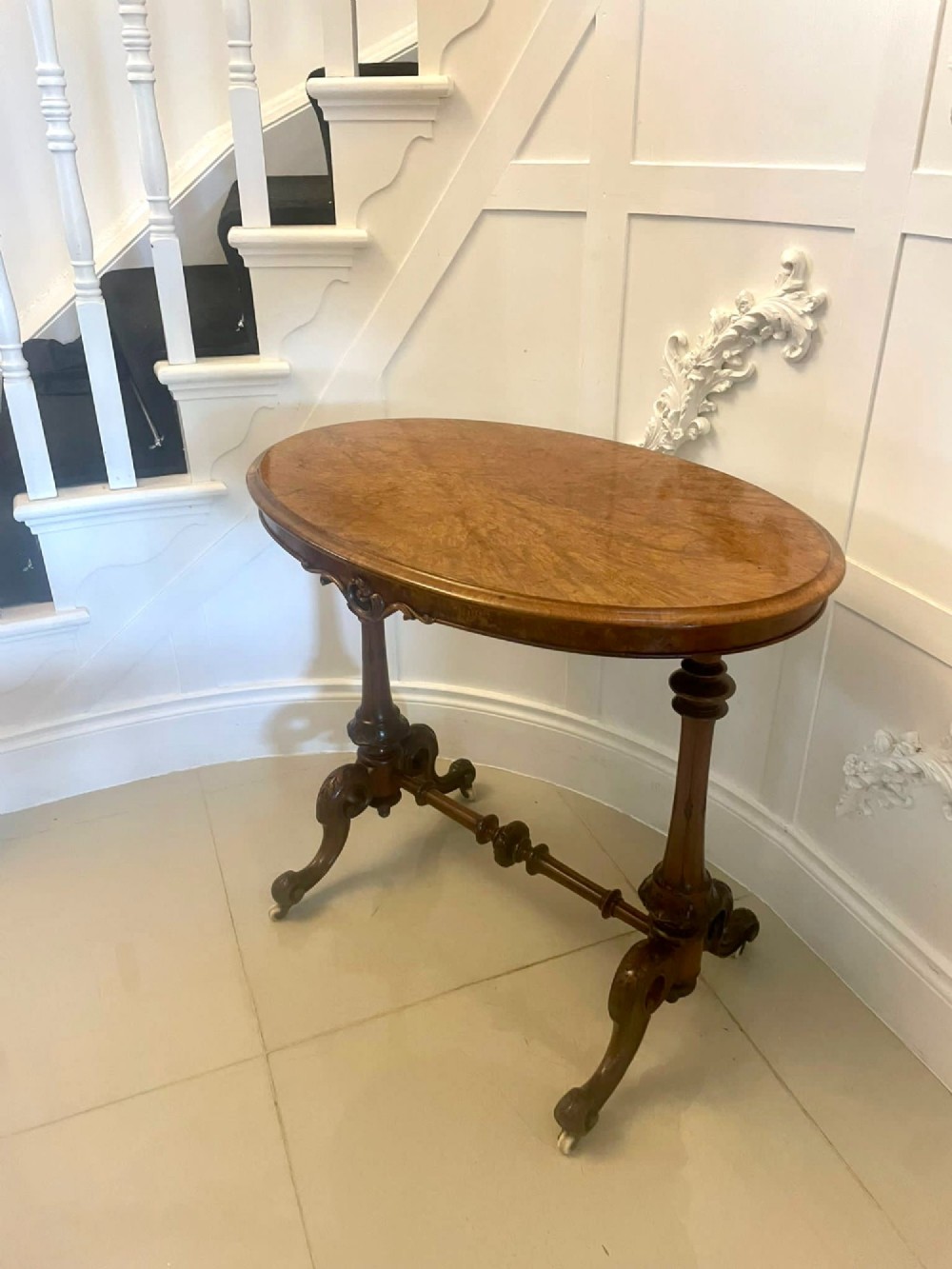 An antique Victorian oval lamp table crafted from burr walnut with turned end supports, a central stretcher, and carved legs with castors. View XIV