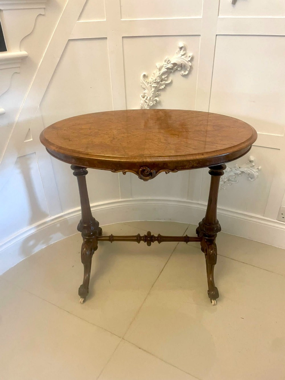 An antique Victorian oval lamp table crafted from burr walnut with turned end supports, a central stretcher, and carved legs with castors. View XV