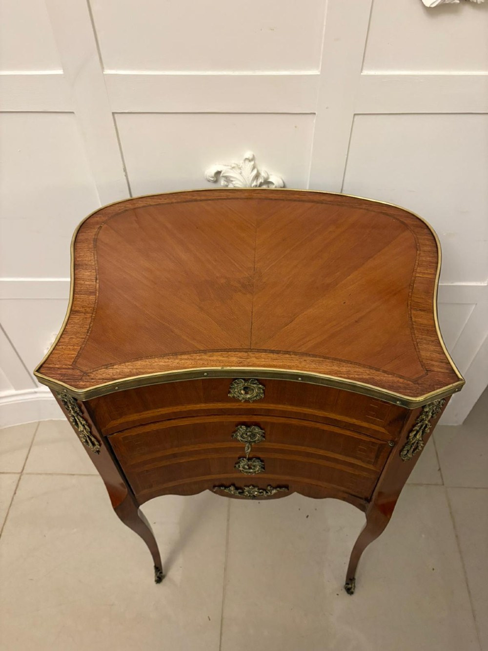 An antique French Louis XV style wooden side cabinet featuring three marquetry drawers, brass rim, decorative corner mounts, and slender cabriole legs. View IV
