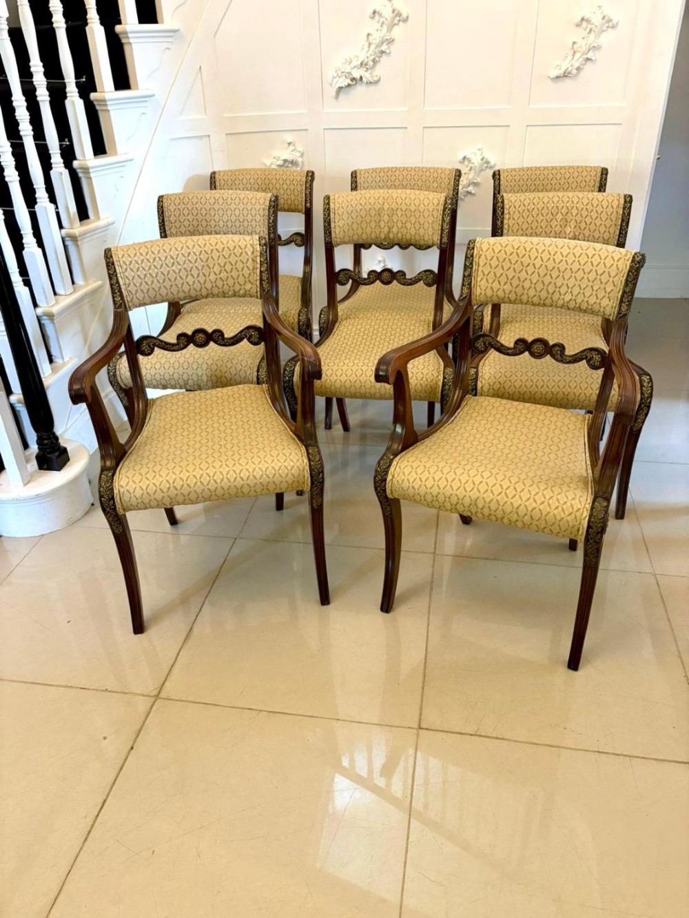 A set of eight antique Regency mahogany dining chairs featuring patterned gold upholstery, carved scrolling mid-rails, and brass detailing.