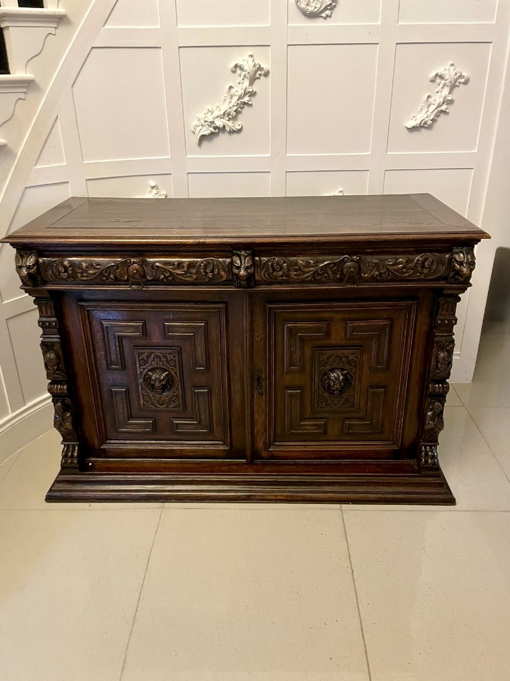 A dark carved oak sideboard featuring geometric panelled cupboard doors with central lion mask carvings and a detailed relief frieze.