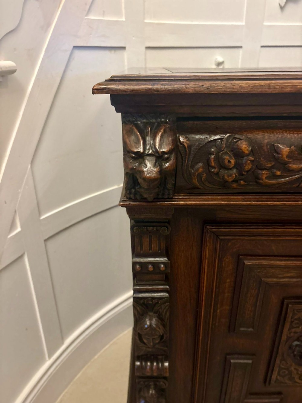 A dark carved oak sideboard featuring geometric panelled cupboard doors with central lion mask carvings and a detailed relief frieze. View II