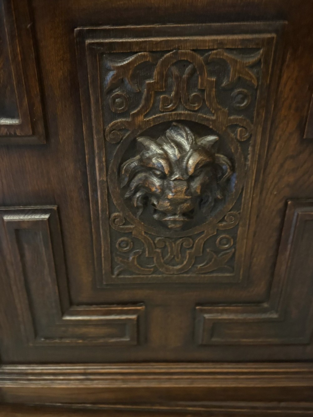 A dark carved oak sideboard featuring geometric panelled cupboard doors with central lion mask carvings and a detailed relief frieze. View IV
