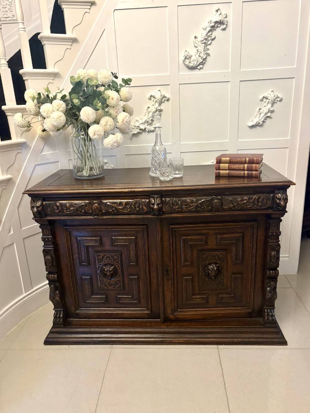 A dark carved oak sideboard featuring geometric panelled cupboard doors with central lion mask carvings and a detailed relief frieze. View VII