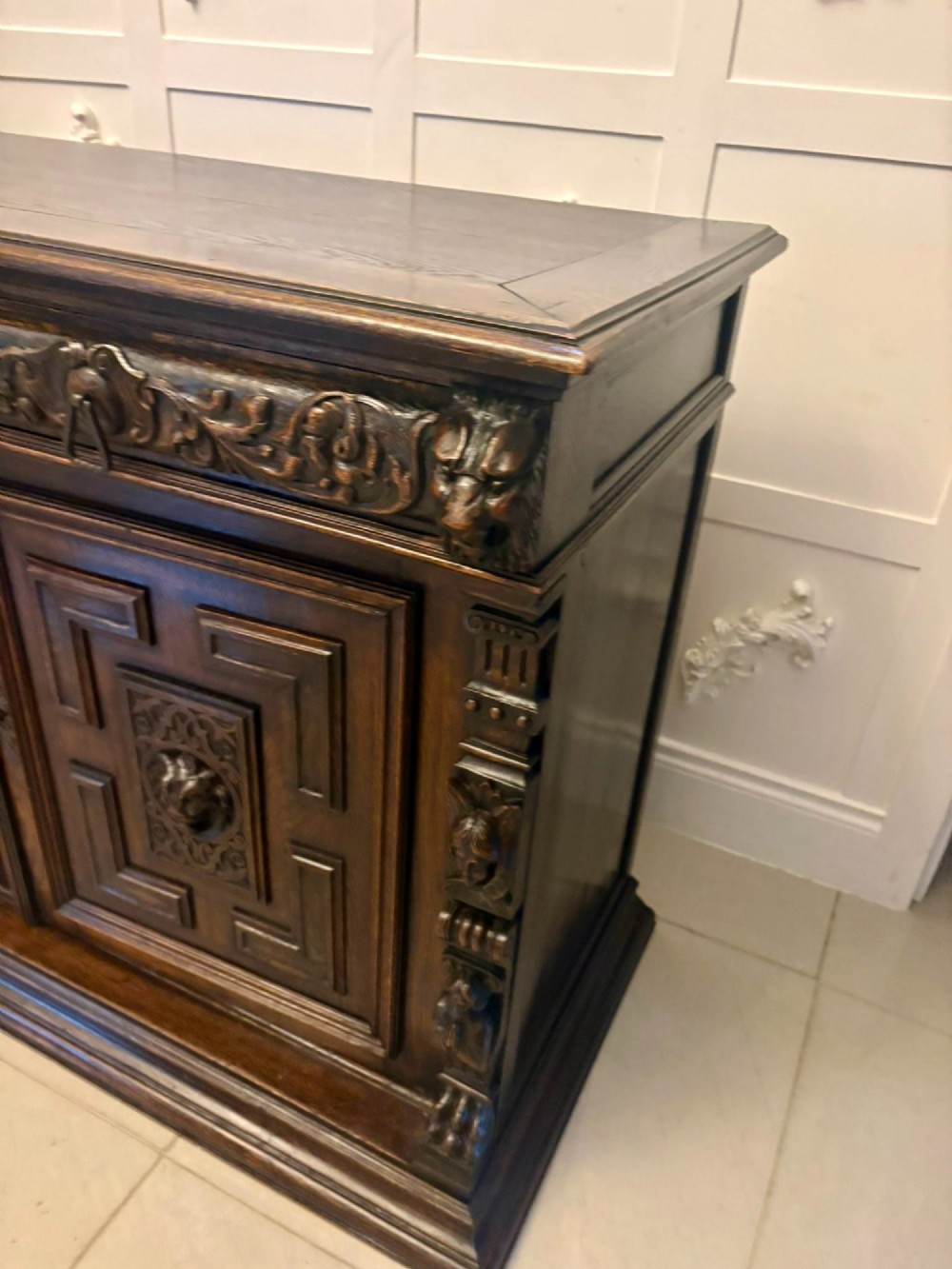 A dark carved oak sideboard featuring geometric panelled cupboard doors with central lion mask carvings and a detailed relief frieze. View XI
