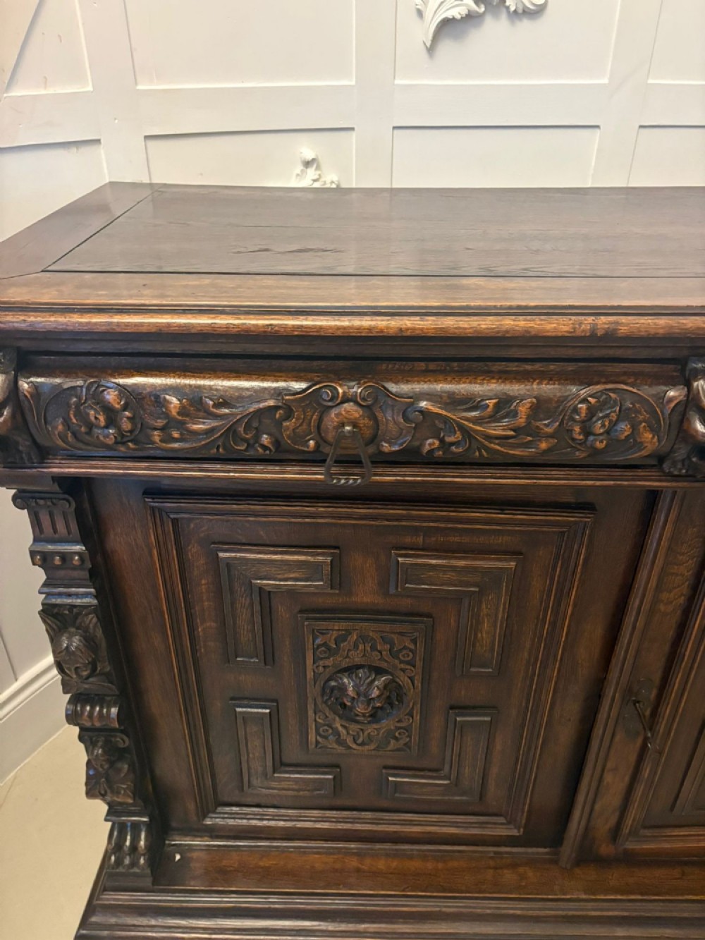 A dark carved oak sideboard featuring geometric panelled cupboard doors with central lion mask carvings and a detailed relief frieze. View XII