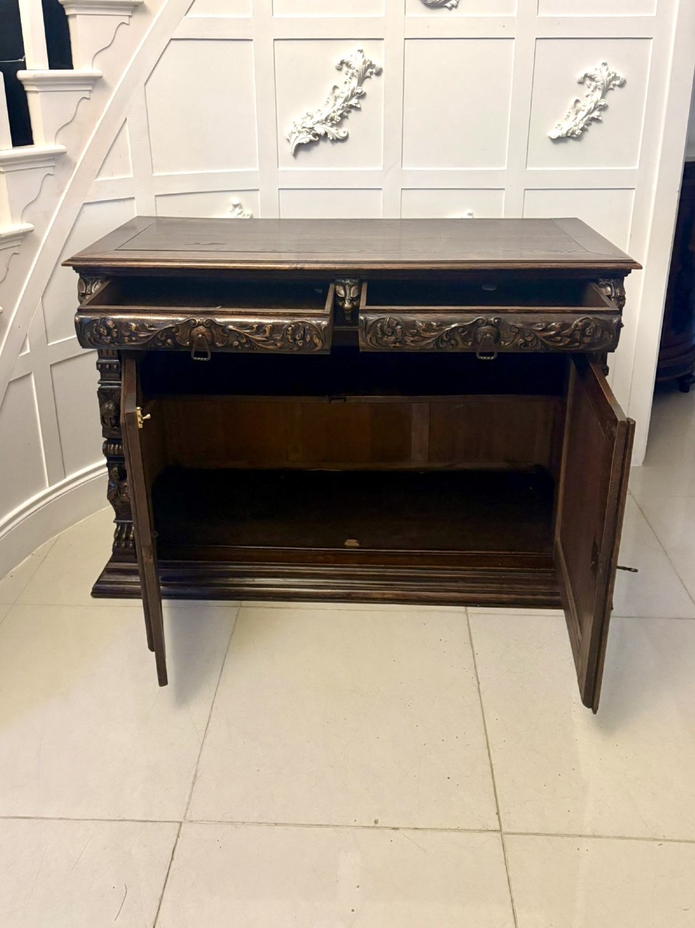A dark carved oak sideboard featuring geometric panelled cupboard doors with central lion mask carvings and a detailed relief frieze. View XIII