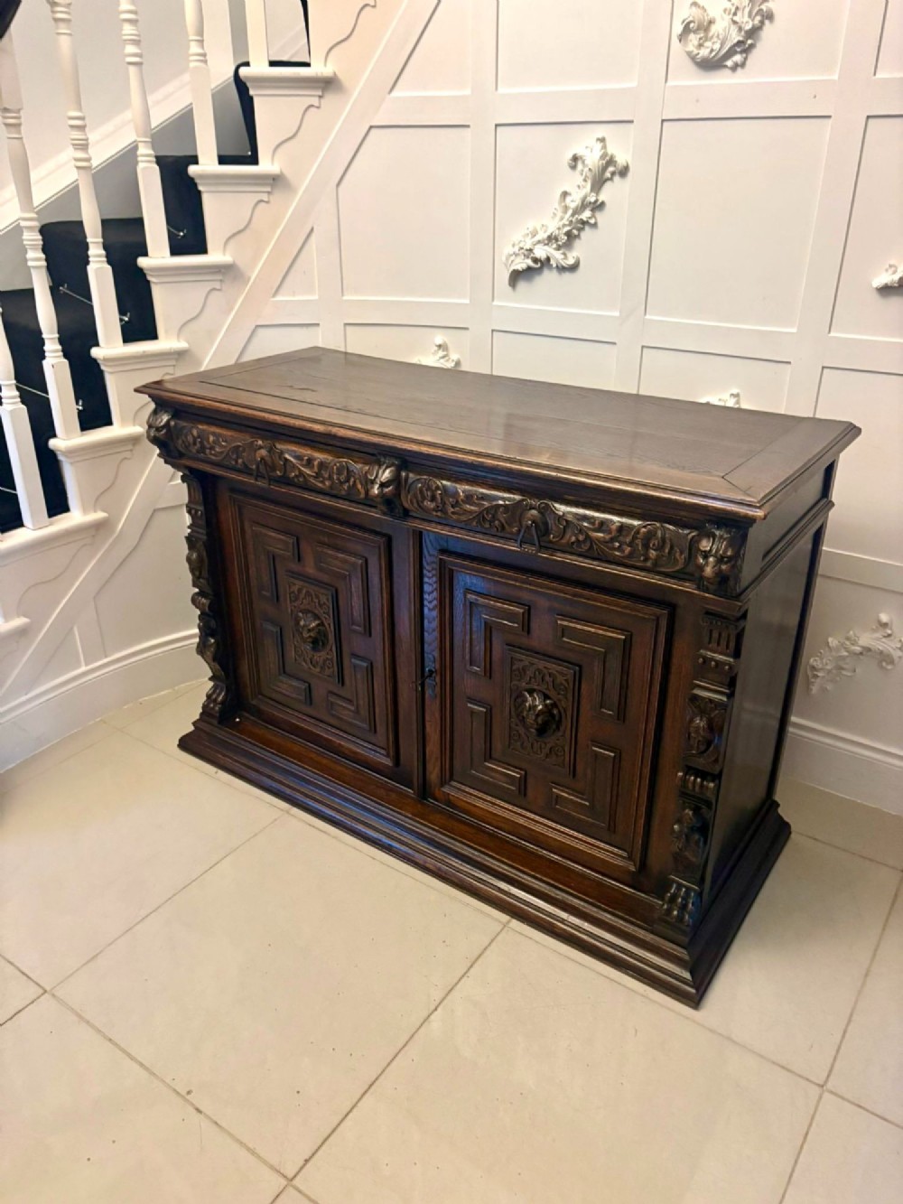 A dark carved oak sideboard featuring geometric panelled cupboard doors with central lion mask carvings and a detailed relief frieze. View XIV