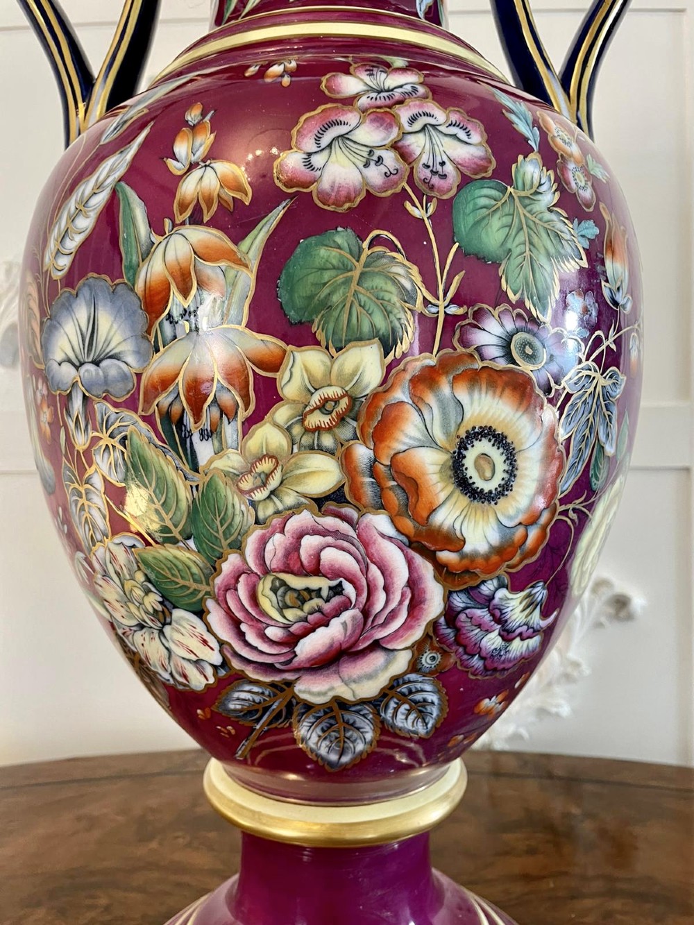 A large Victorian twin-handled porcelain urn vase in vibrant magenta featuring hand-painted botanical florals, navy blue handles, and gold leaf gilding. View II