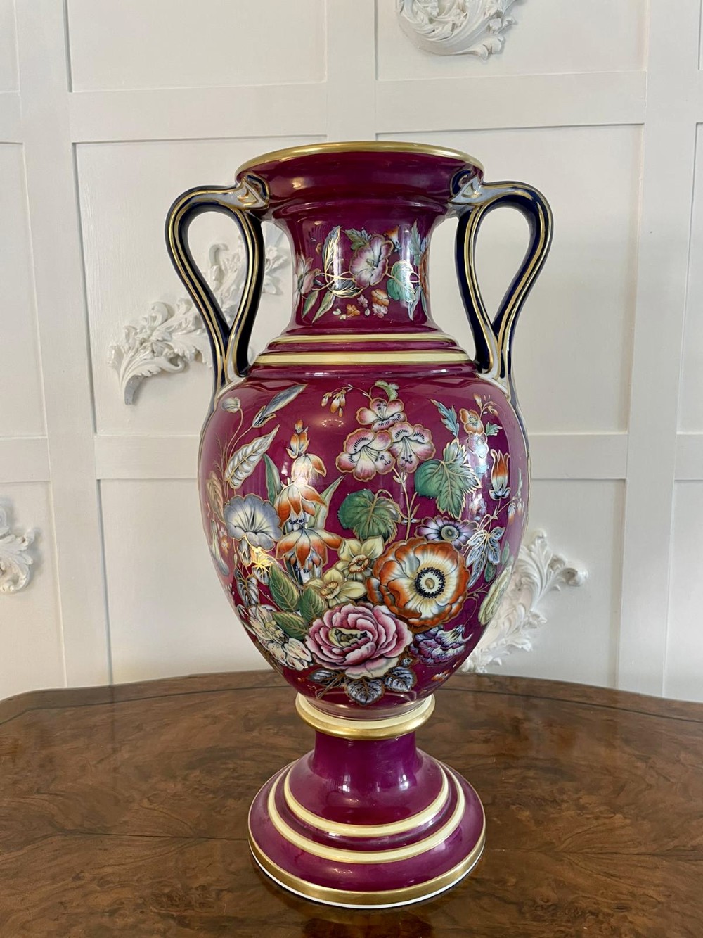 A large Victorian twin-handled porcelain urn vase in vibrant magenta featuring hand-painted botanical florals, navy blue handles, and gold leaf gilding. View V