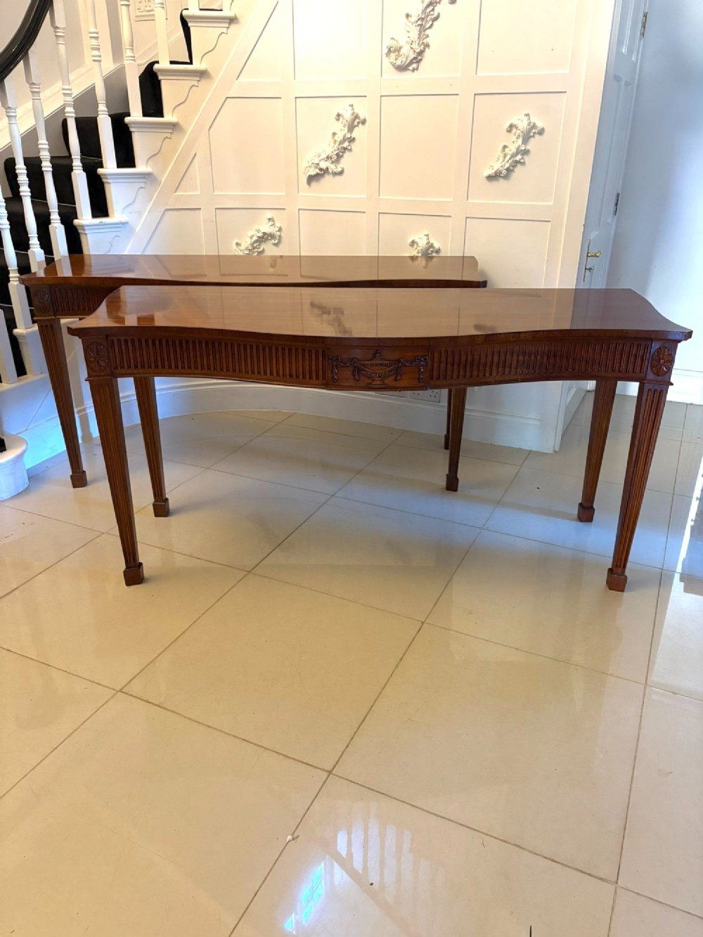 A pair of polished mahogany serpentine-front console tables featuring fluted aprons, carved central urn motifs, and tapered fluted legs. View IV