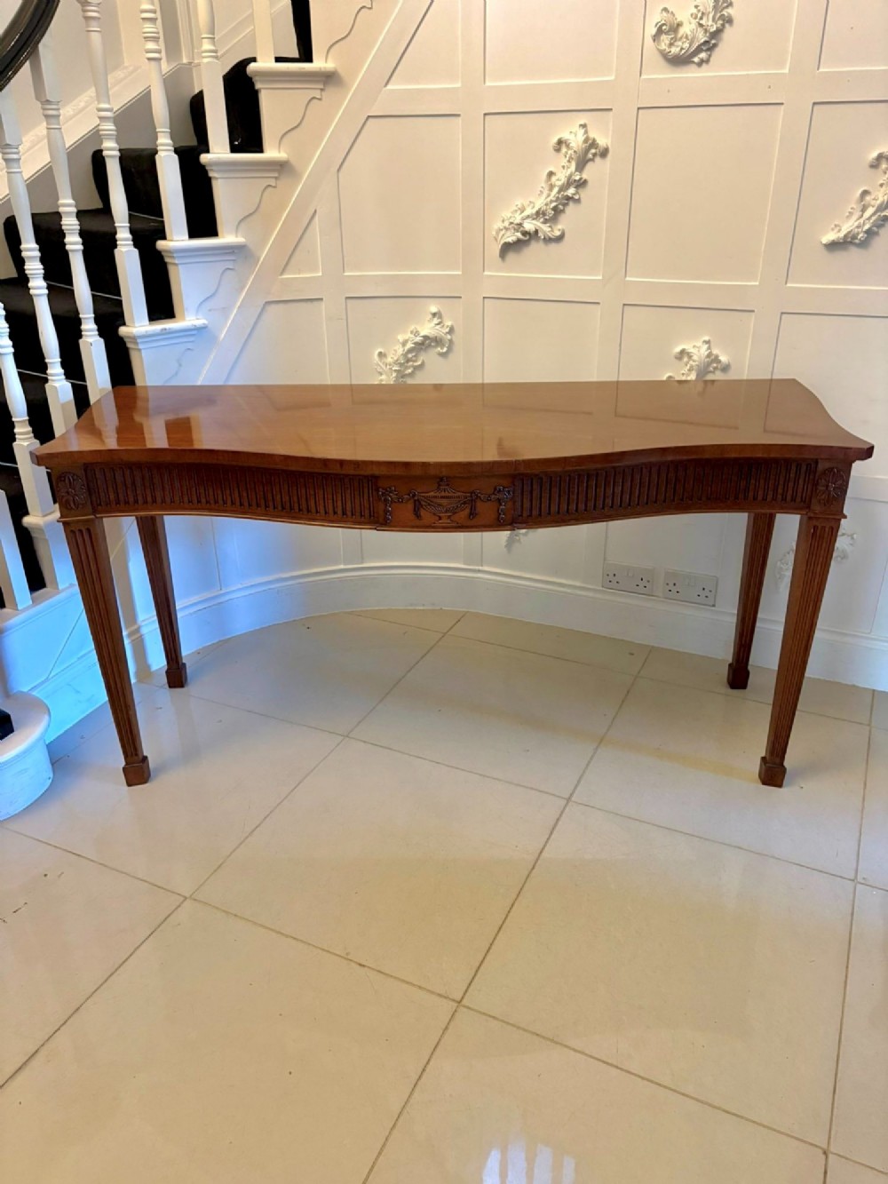 A pair of polished mahogany serpentine-front console tables featuring fluted aprons, carved central urn motifs, and tapered fluted legs. View IX