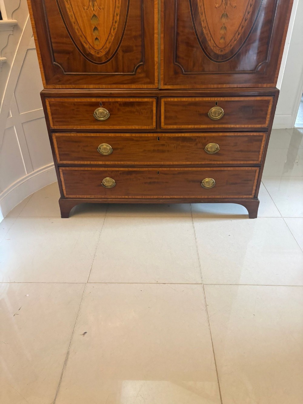 An antique Georgian mahogany linen press featuring two doors decorated with oval satinwood urn marquetry inlays situated above four drawers with brass handles. View V
