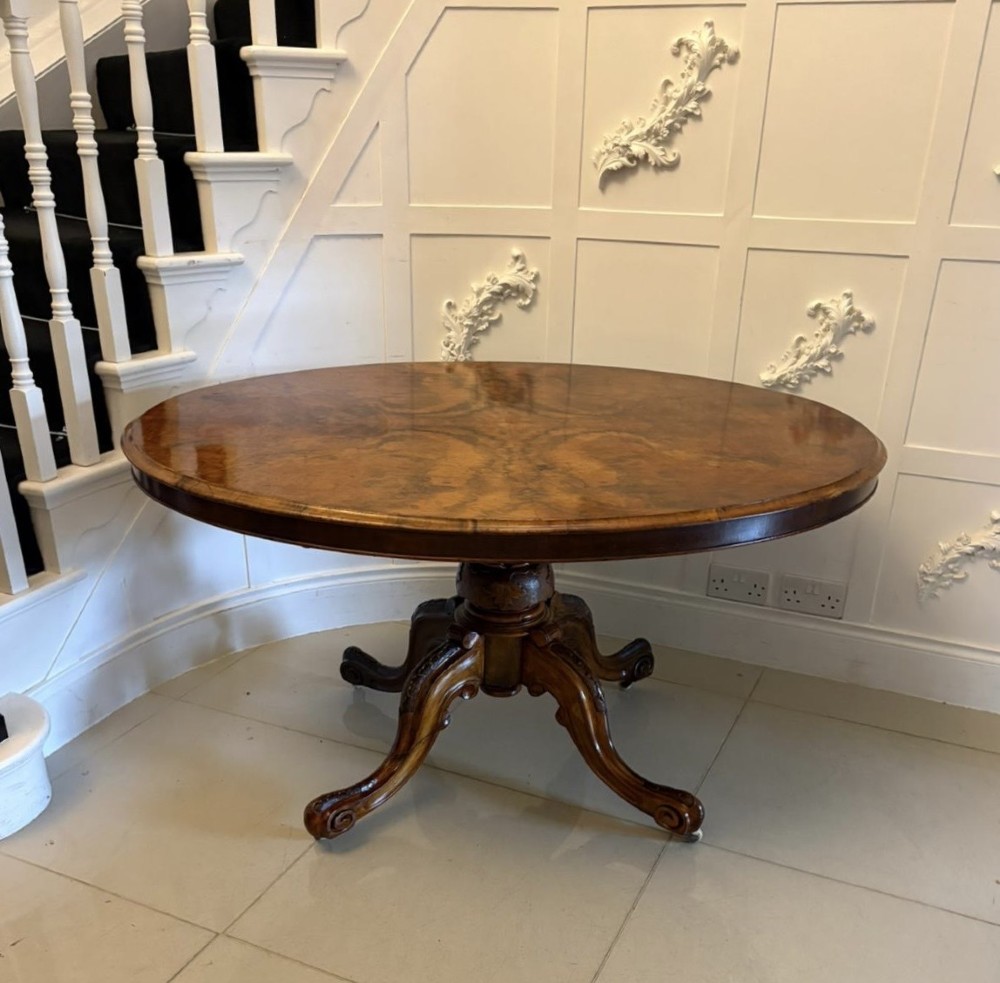 An antique Victorian oval tilt-top dining table featuring book-matched burr walnut grain mounted on a carved central column and tripod base with castors. View VII