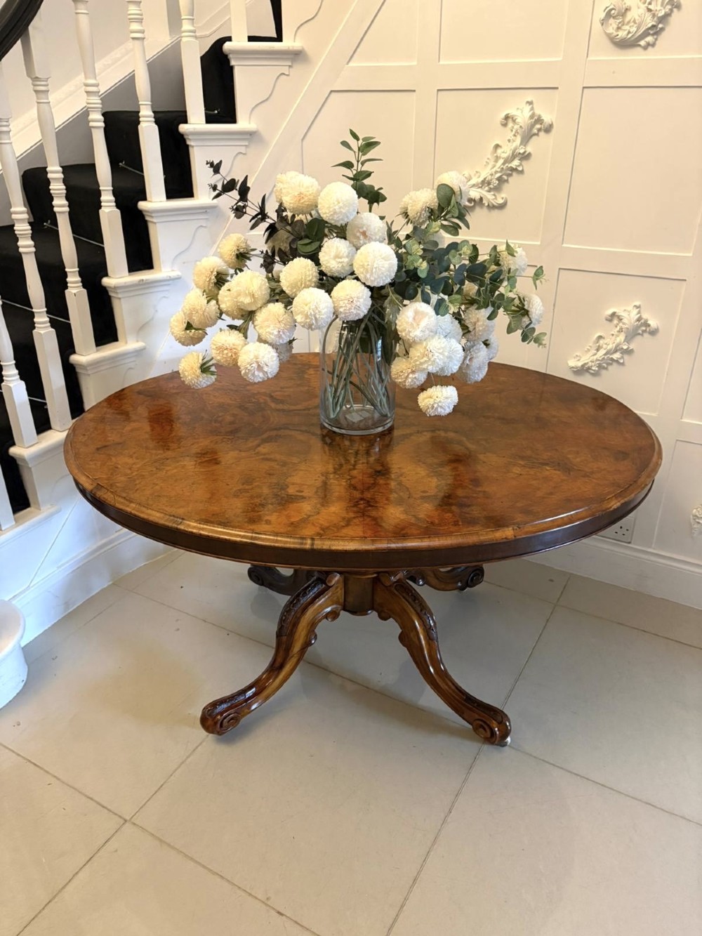 An antique Victorian oval tilt-top dining table featuring book-matched burr walnut grain mounted on a carved central column and tripod base with castors. View VIII