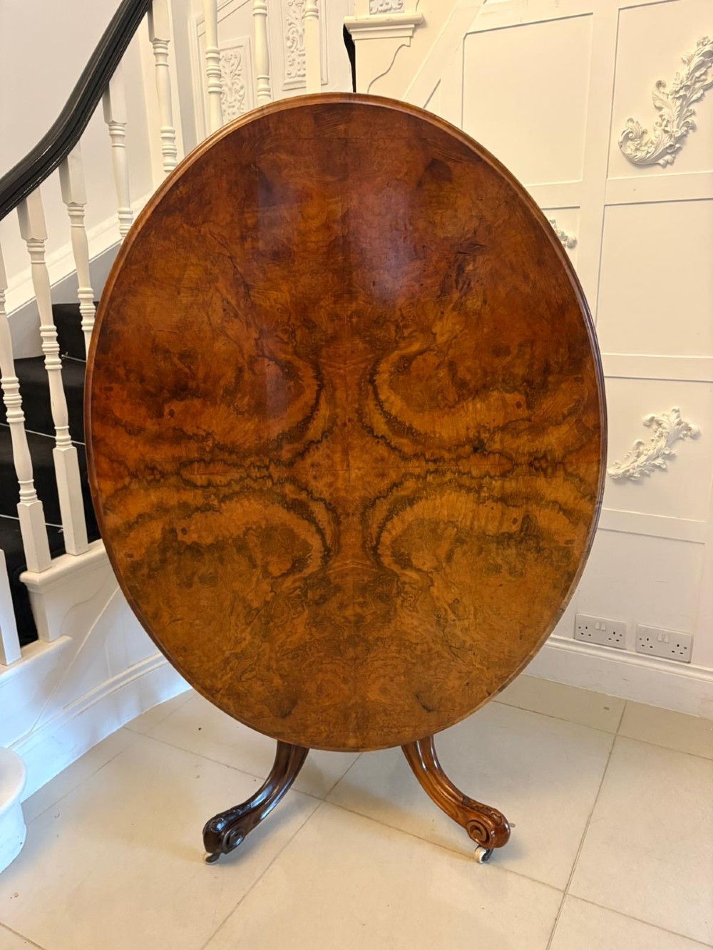 An antique Victorian oval tilt-top dining table featuring book-matched burr walnut grain mounted on a carved central column and tripod base with castors. View XI