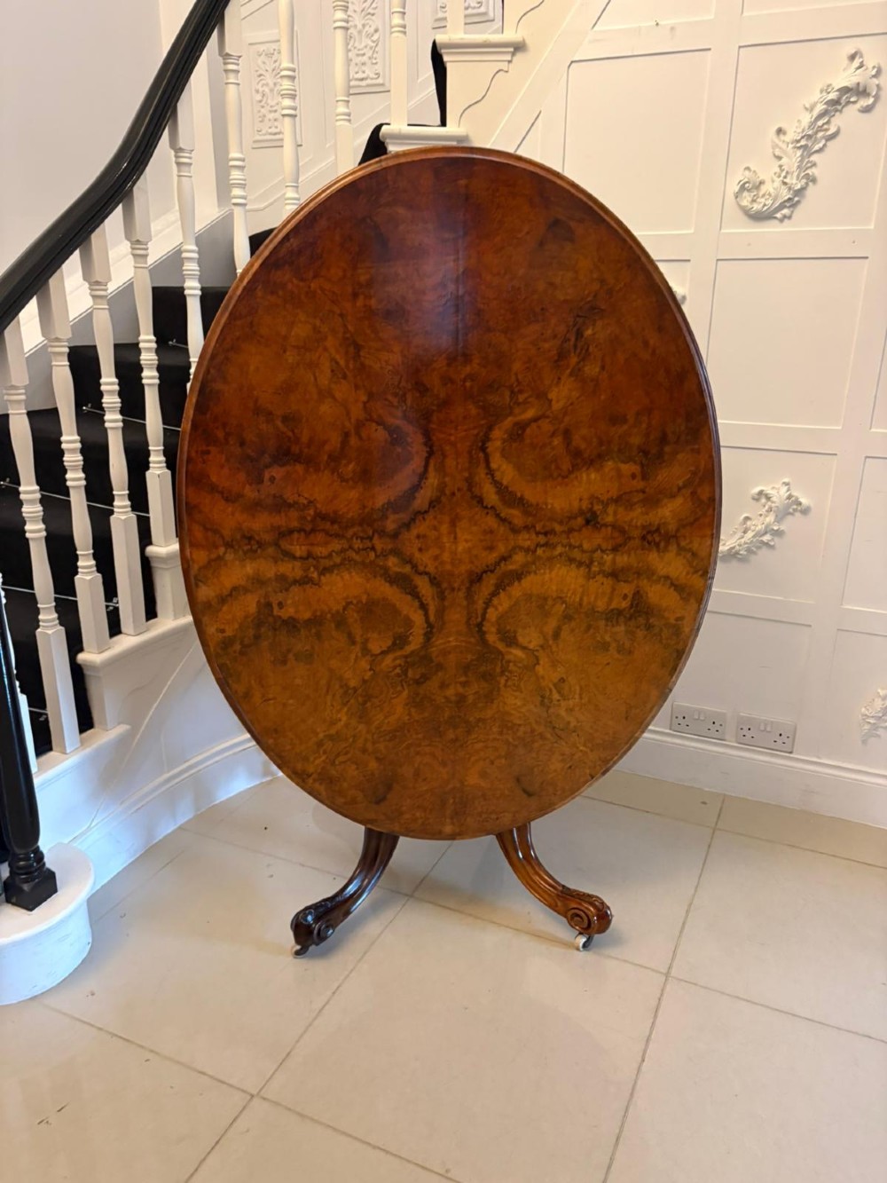 An antique Victorian oval tilt-top dining table featuring book-matched burr walnut grain mounted on a carved central column and tripod base with castors.