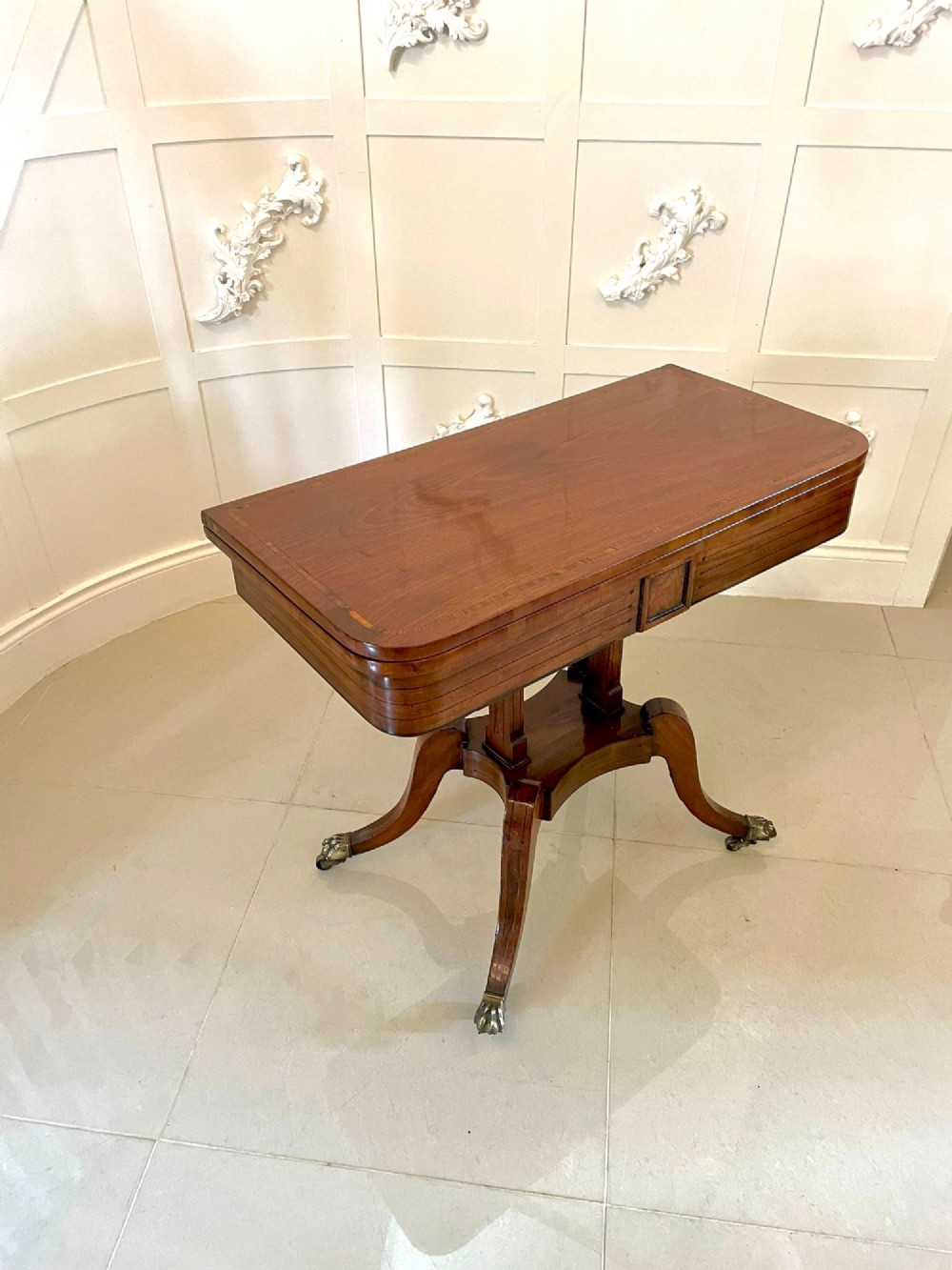 A Regency mahogany fold-over card table featuring an inlaid apron, central pedestal support, and four sabre legs with brass lion paw feet and castors.