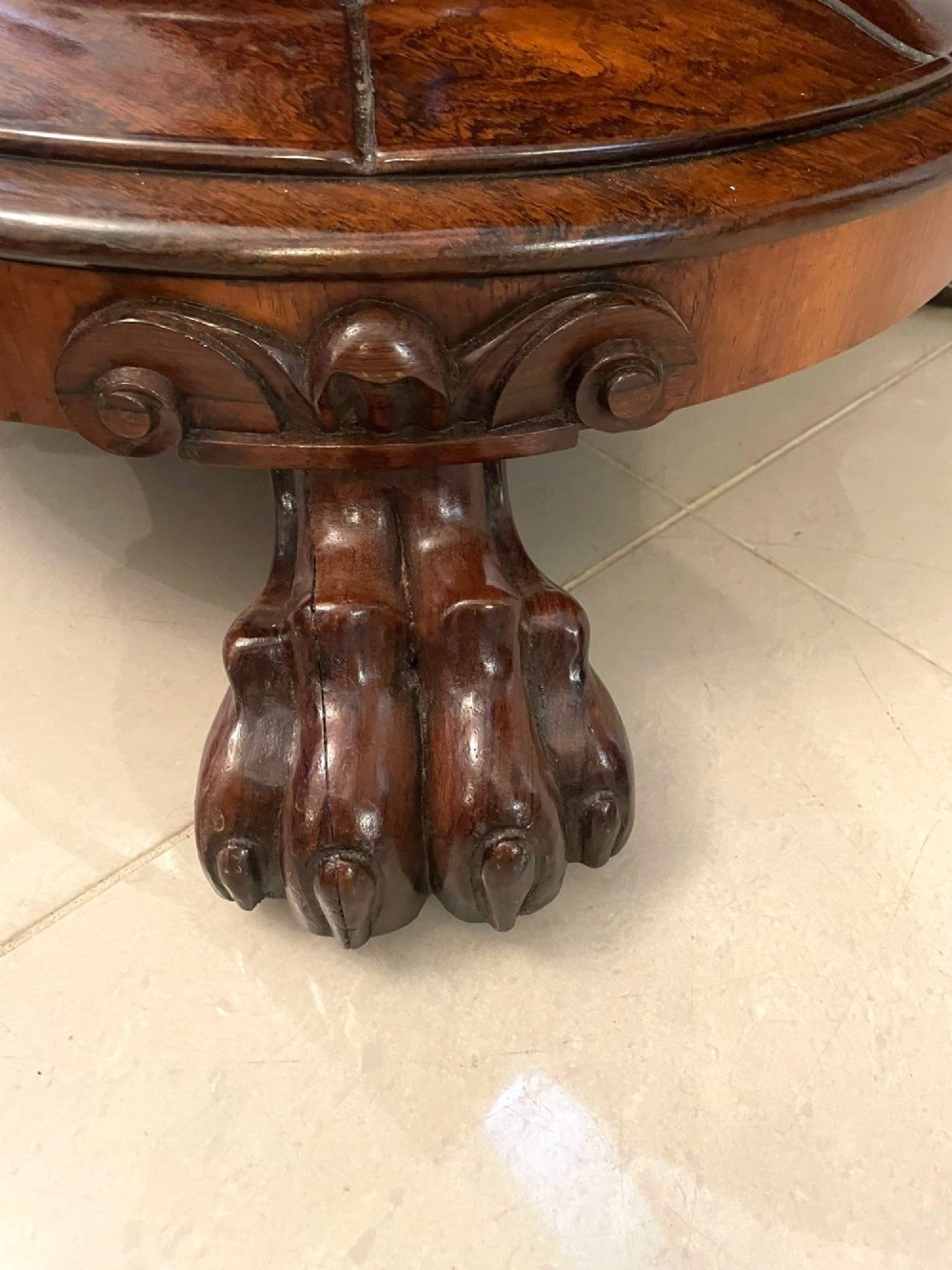 A circular William IV rosewood dining table shown with its top tilted vertically to display the rich wood grain and supported on a carved base with paw feet. View V