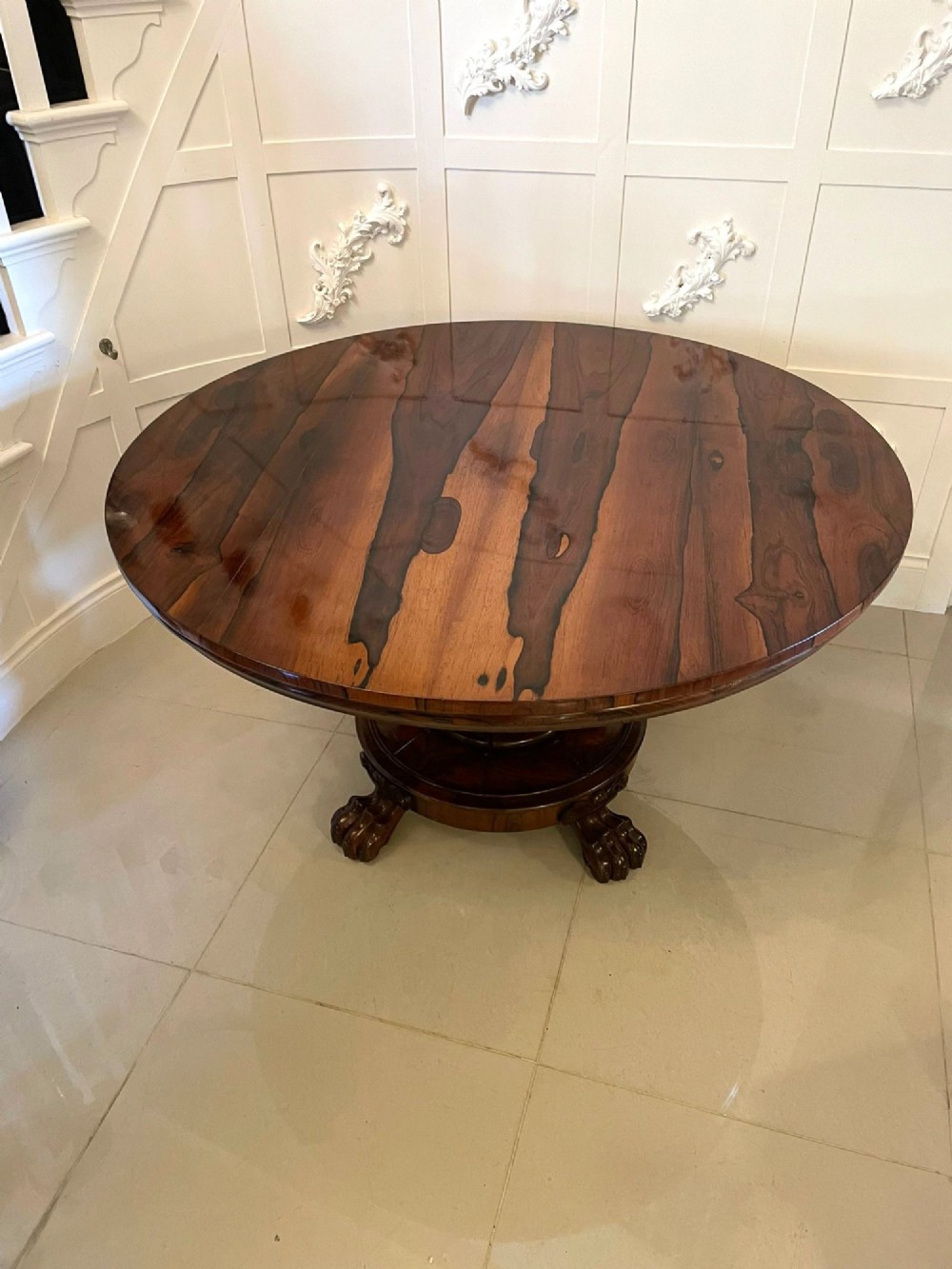 A circular William IV rosewood dining table shown with its top tilted vertically to display the rich wood grain and supported on a carved base with paw feet. View VI