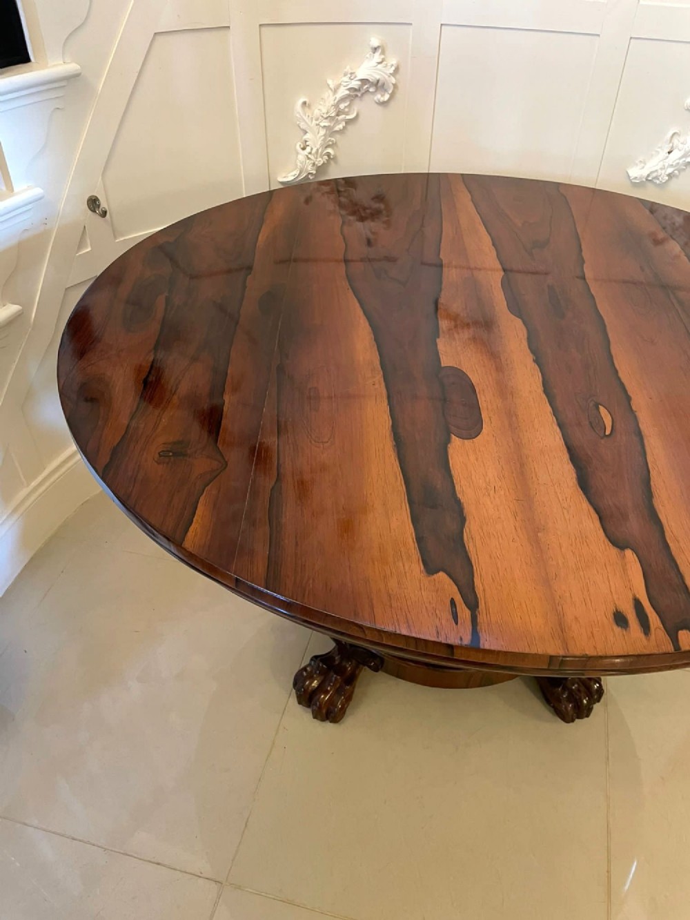 A circular William IV rosewood dining table shown with its top tilted vertically to display the rich wood grain and supported on a carved base with paw feet. View XII