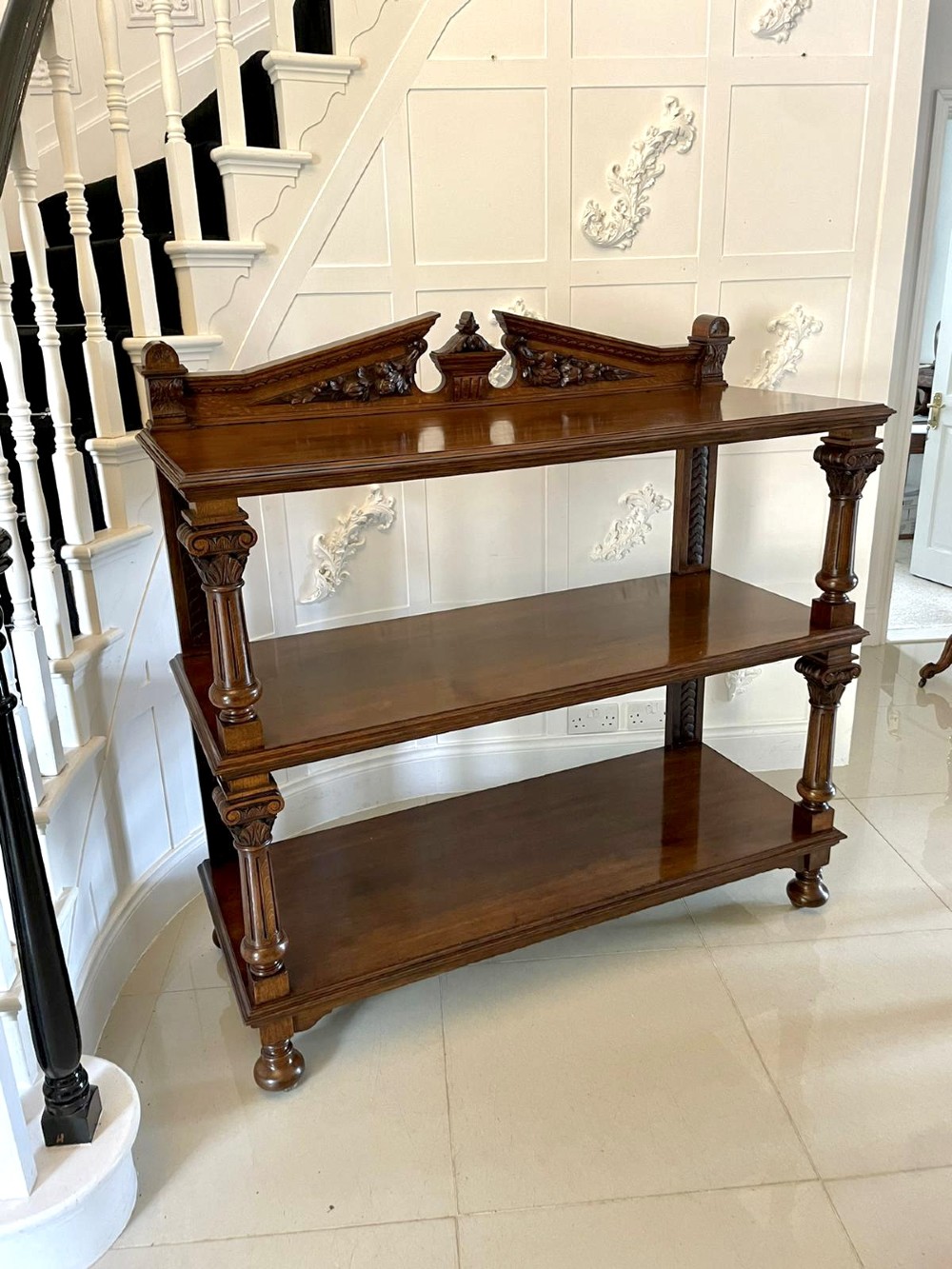 A three-tiered dark oak antique dumbwaiter featuring carved column supports, a carved gallery cresting, and bun feet.