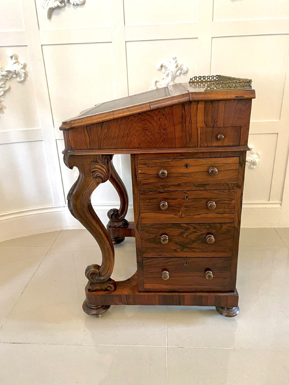 A Victorian rosewood Davenport desk featuring a sloped dark green leather writing surface, decorative brass gallery rail, carved cabriole front supports, and side drawers. View IX