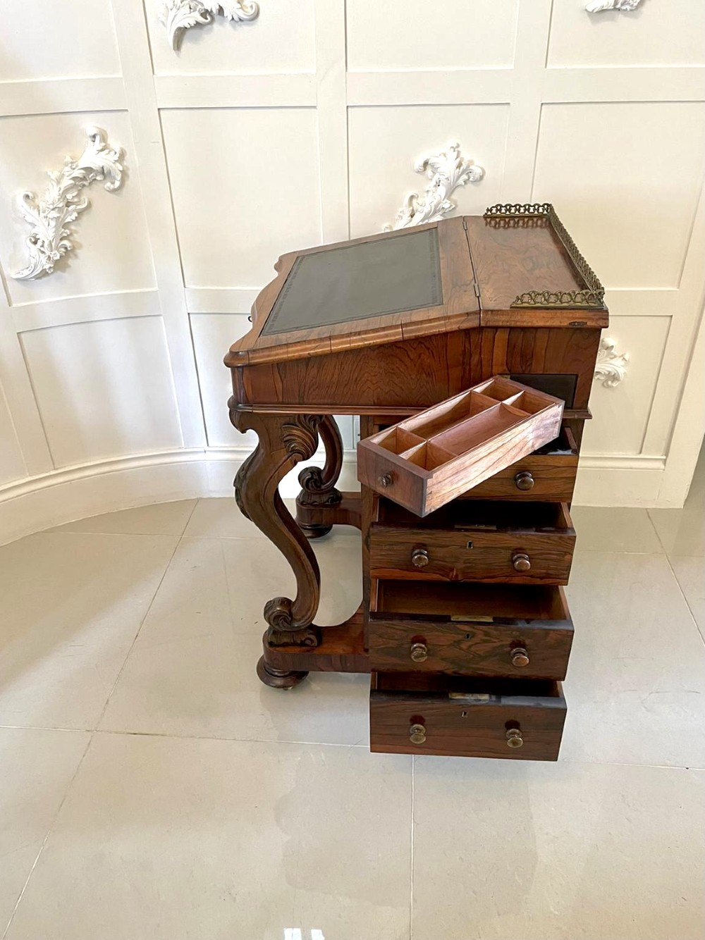 A Victorian rosewood Davenport desk featuring a sloped dark green leather writing surface, decorative brass gallery rail, carved cabriole front supports, and side drawers. View X