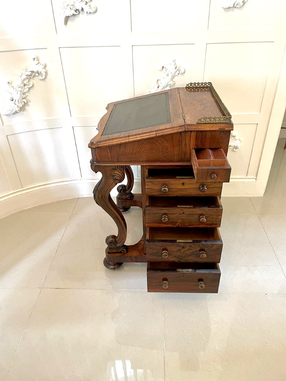 A Victorian rosewood Davenport desk featuring a sloped dark green leather writing surface, decorative brass gallery rail, carved cabriole front supports, and side drawers. View XIV