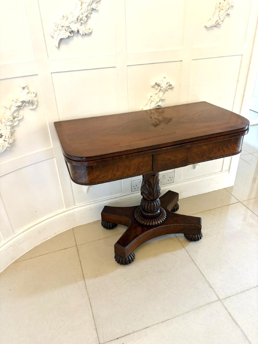A 19th-century William IV figured mahogany card table with a fold-over top, carved central pedestal, and a concave platform base supported by carved feet.