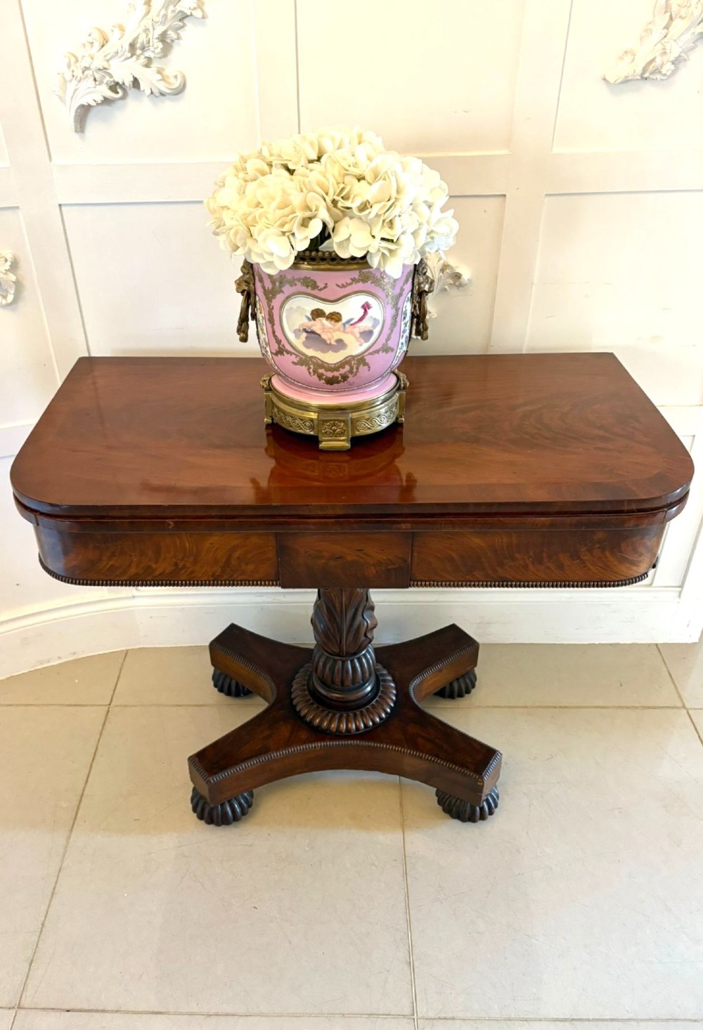 A 19th-century William IV figured mahogany card table with a fold-over top, carved central pedestal, and a concave platform base supported by carved feet. View II