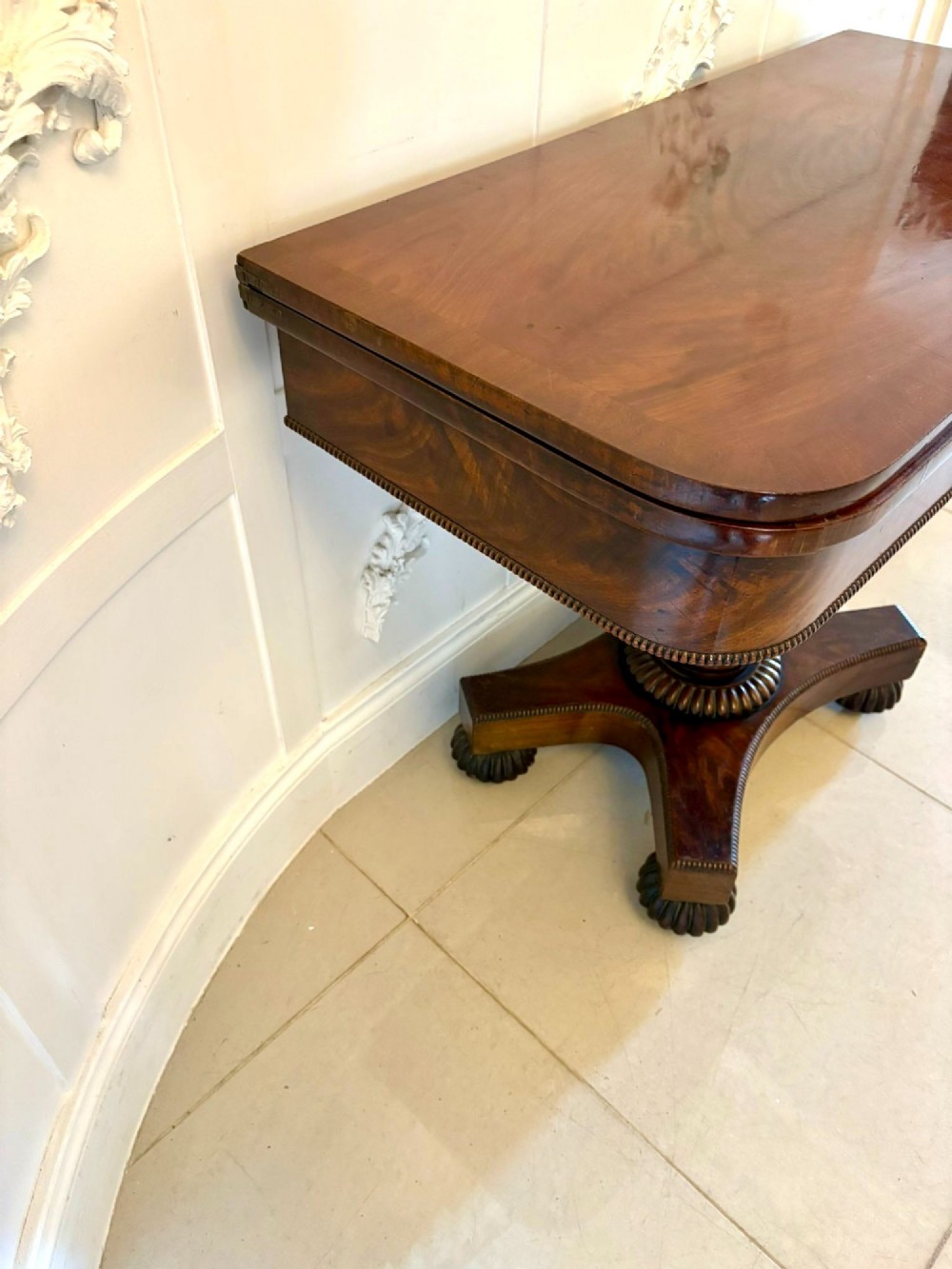 A 19th-century William IV figured mahogany card table with a fold-over top, carved central pedestal, and a concave platform base supported by carved feet. View VI