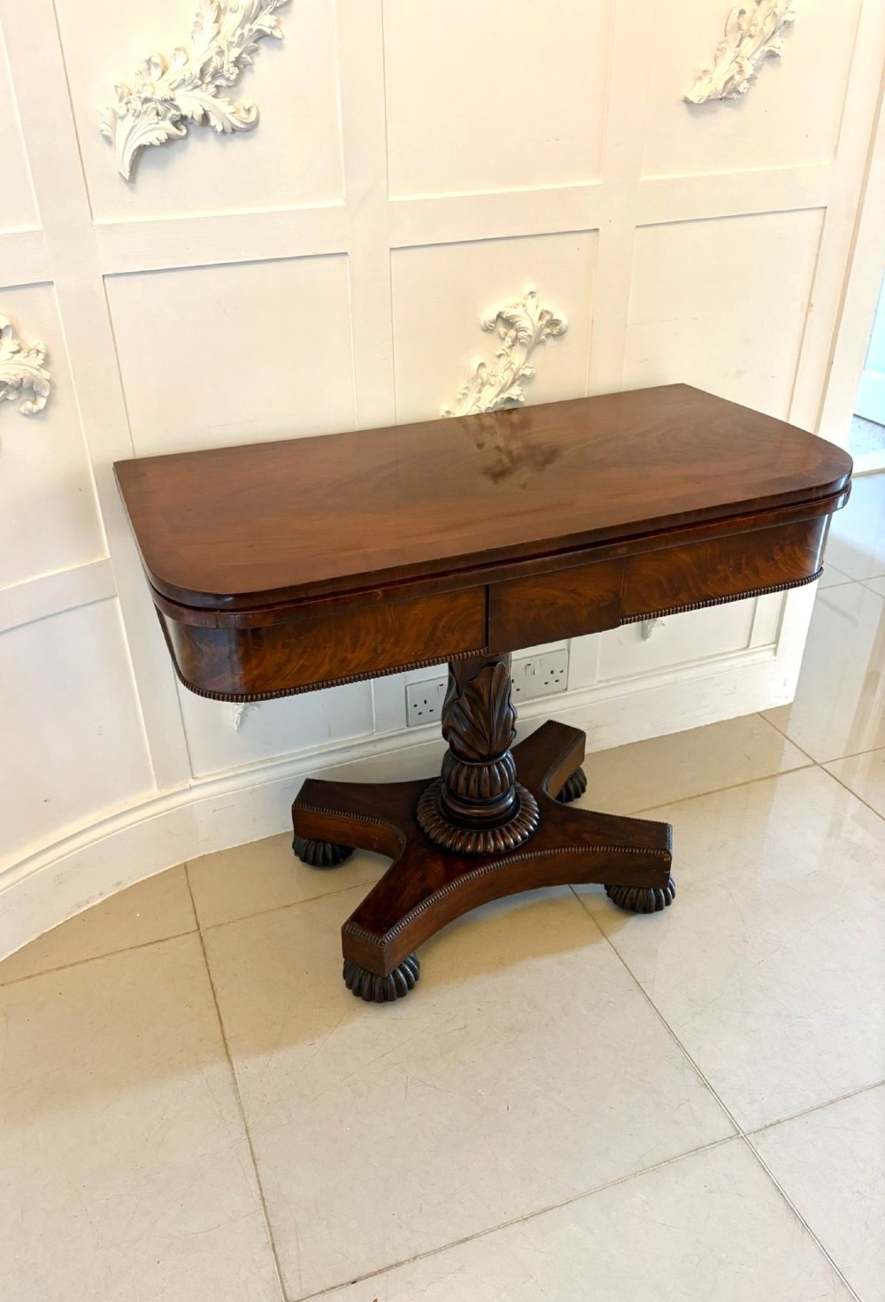 A 19th-century William IV figured mahogany card table with a fold-over top, carved central pedestal, and a concave platform base supported by carved feet. View X